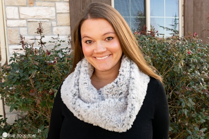 Smiling female model wearing a black sweater and a Kuma colorway example of the Fur-Ever Fur Infinity Scarf Crochet Pattern in a front yard setting.